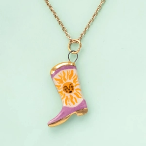Close-up of a white cowboy boot pendant with purple trim and yellow sun face on a gold chain