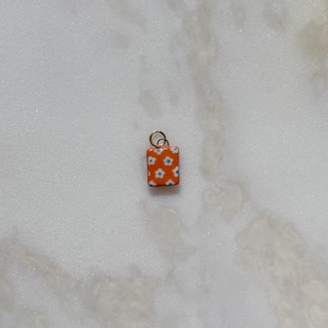 Detalle de dije cuadrado de esmalte naranja con motivo floral blanco y aro dorado