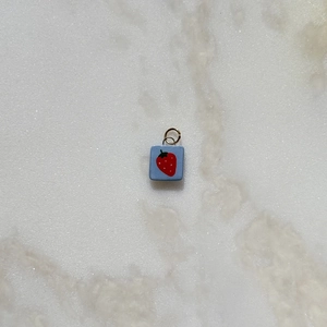 Detalle de un colgante cuadrado azul con motivo de fresa roja y anillo de metal