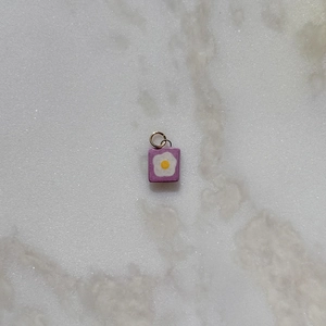 Vista detallada de un colgante rectangular morado con diseño de huevo frito y aro metálico