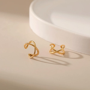 Close-up of minimalist X-shaped gold-plated ear cuffs with sleek mirror finish on beige surface