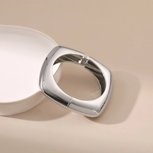 Polished silver square bangle with integrated hinge clasp on a ceramic display tray