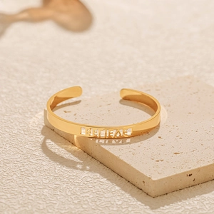 Detail eines goldenen Armreifs mit ausgeschnittenem „BELIEVE“-Wort auf strukturiertem Stein