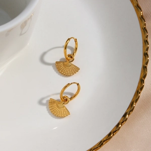 Detail shot of golden fan-shaped stainless steel earrings on a decorative white porcelain tray