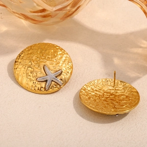 Golden textured shield stud earrings with silver starfish detail on a sandy backdrop