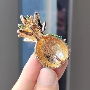 Broche en forme d'ananas doré avec de l'émail vert tenue par une main de femme