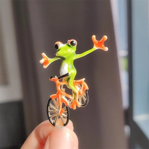 Detail of vibrant green frog riding an orange bicycle enamel brooch held in hand