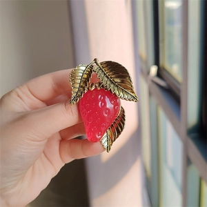 Macro detail of red resin strawberry brooch with golden metal leaves held in hand