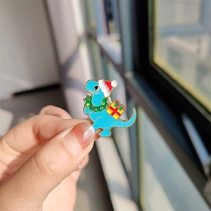 Détail d'un pin's dinosaure de Noël bleu avec bonnet de Père Noël et cadeau sur une main
