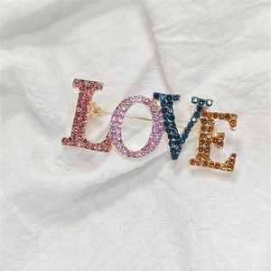 Goldene LOVE Buchstaben-Brosche mit bunten Strasssteinen auf weißem Stoffhintergrund