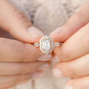 Rose gold emerald cut halo engagement ring held in hand with pave set accents