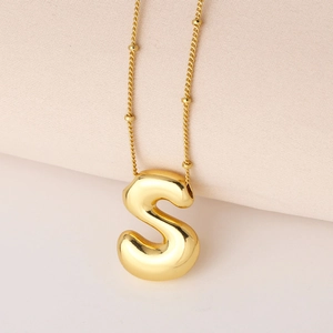 Detail eines polierten goldenen S-Buchstaben-Anhängers mit Perlenkette auf Creme-Stoff