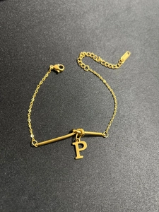 Detail eines Gold-Titan-Armbands mit Buchstaben-P-Anhänger und Metallstab auf dunklem Grund