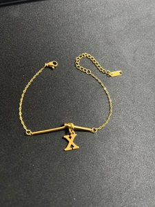Detail eines vergoldeten Edelstahlknoten-Armbands mit Buchstabe-X-Anhänger und Karabiner