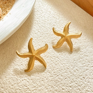 Macro detail of starfish stud earrings with a textured gold finish on a beach sand background