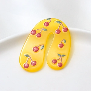 Close-up of yellow U-shaped acrylic earrings with cherry patterns on a white ceramic tray