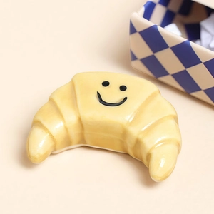 Süßer gelber Croissant-Anhänger mit Smiley-Design auf warmem beigem Hintergrund