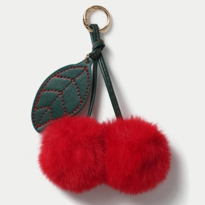Macro detail of plush red cherry bag charm with green leather leaf and gold clasp
