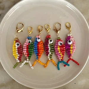 Close-up of six colorful handmade beaded fish pendants with gold lobster clasps on a ceramic dish