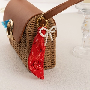 Brown straw handbag with paisley scarf and pearl bow keychain on off-white background