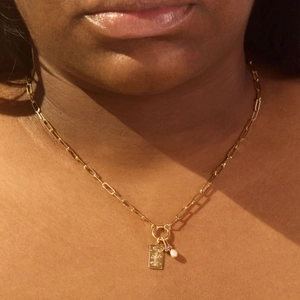 Gold paperclip chain necklace with floral engraved plaque, red gemstone, and pearl charms