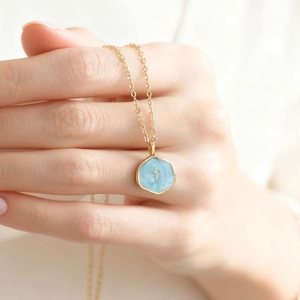 Close-up of golden cross necklace with light blue hexagonal enamel pendant held by hand