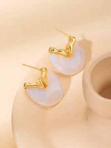 Close-up of golden irregular earrings with pearlescent white resin pendant on beige background