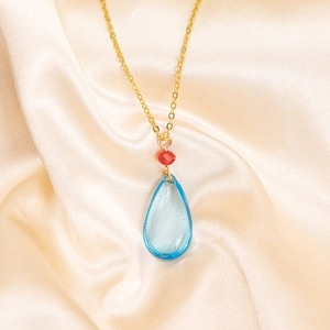 Close-up of golden necklace with red bead and light blue teardrop pendant on satin