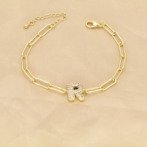 Macro detail of gold paperclip chain bracelet with a sparkling crystal letter R charm