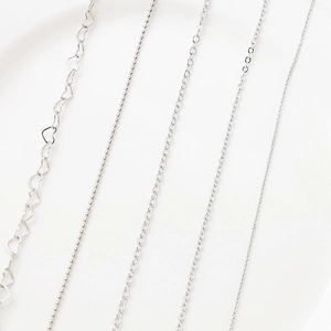 Cinq styles de chaînes de bijoux DIY argentées avec maillons cœur et perles sur blanc