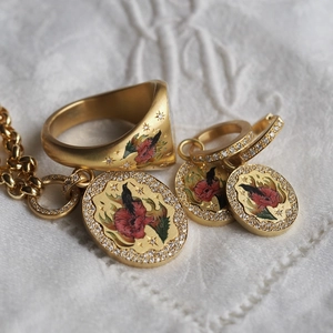 Detalle de set de joyería dorada con esmalte de flores rojas y ave sobre tela blanca