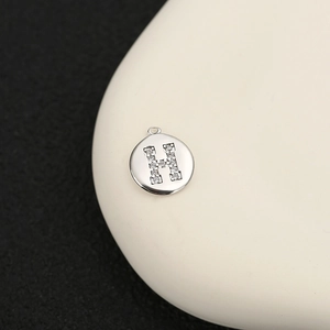 Detalle de un colgante redondo plateado con circonitas y la letra H para joyería DIY