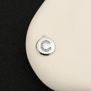 Vista macro de un colgante circular de plata con letra C y circonitas sobre piedra blanca