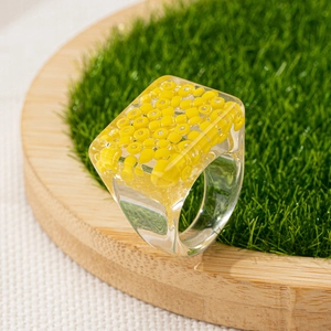 Detail of a transparent resin square ring featuring bright yellow beads on green turf