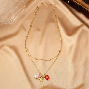 Detalle de collar dorado de dos capas con llave corazón, perla y circón rojo