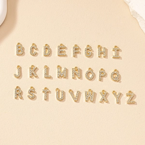 Set di ciondoli con lettere dorate e cristalli pavé su superficie beige testurizzata