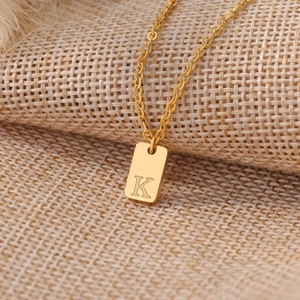 Gold rectangular pendant necklace with engraved letter K on a textured linen background