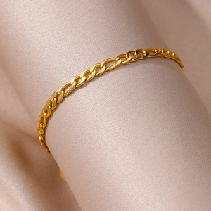 Close-up of golden Figaro anklet resting on luxurious smooth champagne silk fabric