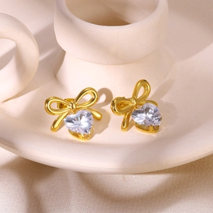 Close-up of golden bow stud earrings with heart-shaped crystal on white ceramic