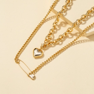 Layered gold necklace featuring heart pendant and safety pin charm on textured stone