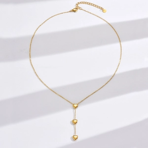 Détail du collier en Y en acier doré avec trois pendentifs en cœur sur un fond blanc épuré