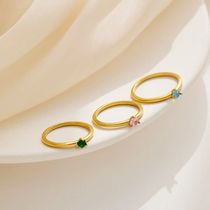 Tres anillos dorados con piedras verdes rosas y azules sobre cerámica blanca y seda