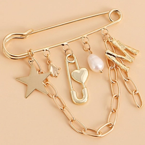 Gold safety pin brooch with star, heart, pearl, and letter M charms on a pink background