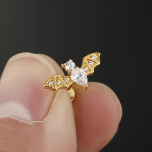 Gold bat-shaped piercing stud with white zircon crystals shown in hand for scale