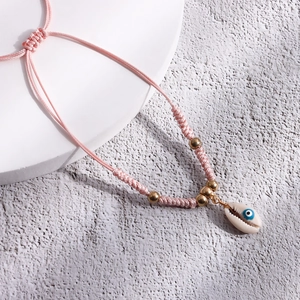 Close-up of pink woven anklet with golden beads, shell charm and blue evil eye detail