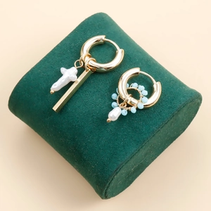 Asymmetric gold hoop earrings with cross pearl and blue bead detail on green velvet