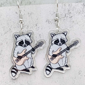 Detalle de pendientes de acrílico con mapache y guitarra eléctrica sobre piedra