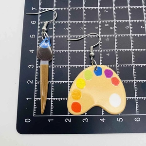 Artistic acrylic earrings featuring a colorful paint palette and paintbrush on a scale mat