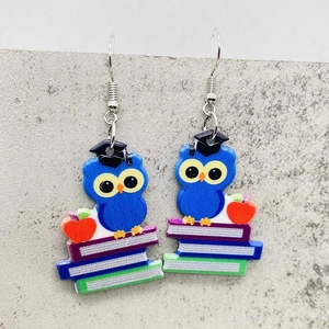 Détail de boucles d'oreilles en acrylique avec chouette sur livres et pomme rouge