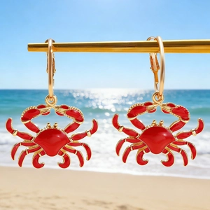 Exaggerated golden crab earrings with red enamel detail on a sunny beach background